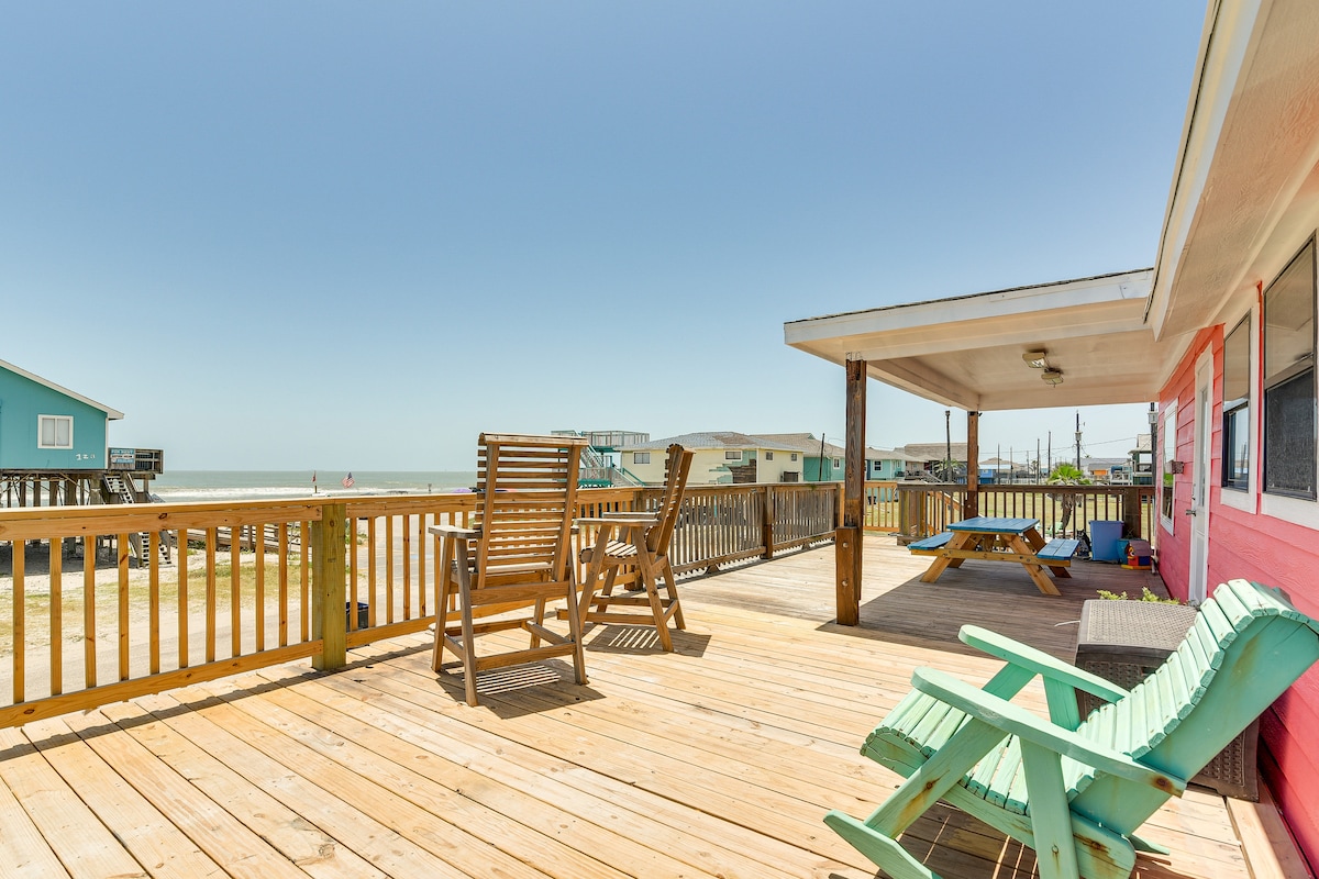 A spacious deck extends from the house, overlooking the beach and ocean. Comfortable seating options are provided, including adirondack chairs and a picnic table, allowing for relaxing outdoor dining. The surrounding area features nearby beach homes, showcasing a coastal atmosphere.