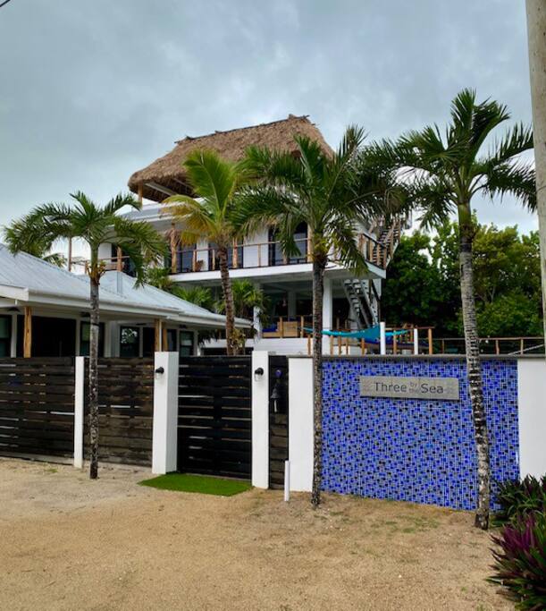 A multi-level property is framed by lush palm trees, with a thatched roof providing a tropical touch. The entrance features a decorative tiled wall displaying the name 'Three to Sea.' A sandy path leads up to the welcoming gated area of the home.