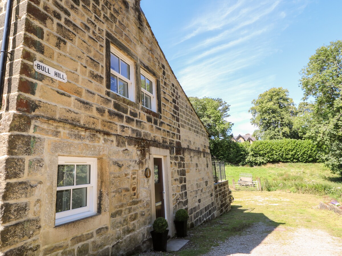 The exterior of Bull Hill Cottage showcases its stone façade, featuring multiple windows that capture sunlight. The entrance is marked by a small round sign displaying 'Bull Hill'. Lush greenery and a well-maintained area are visible in the background, presenting a serene country setting.