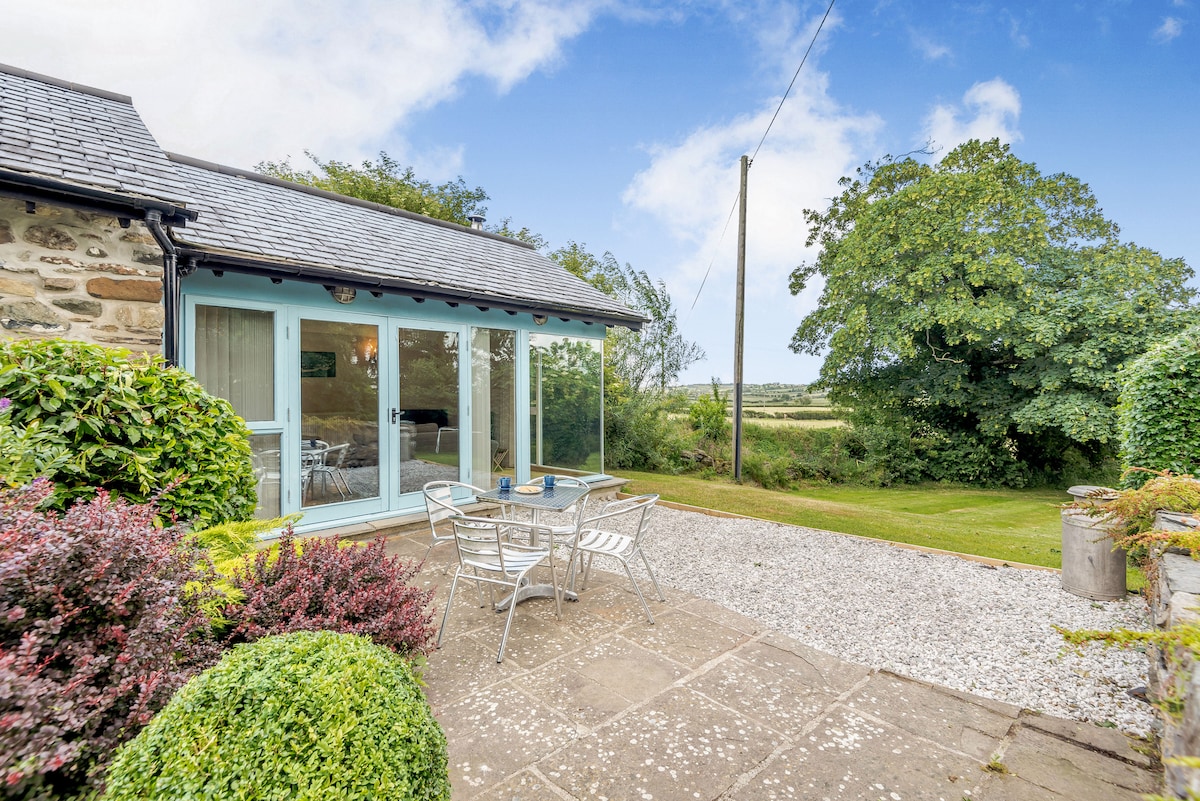 A charming outdoor patio area is seen with metal garden furniture placed on a stone surface, surrounded by neatly trimmed hedges and colorful plants. A lush green lawn extends into the background, framed by trees against a clear blue sky.
