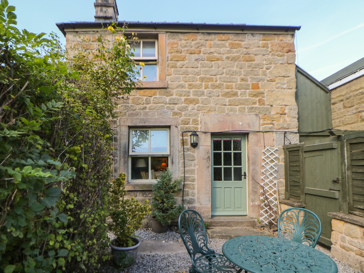 The exterior of Clematis Cottage features stone walls and a welcoming green front door. A small seating area with a round table and decorative chairs is positioned outside, surrounded by lush greenery and garden planters, creating an inviting and peaceful environment.