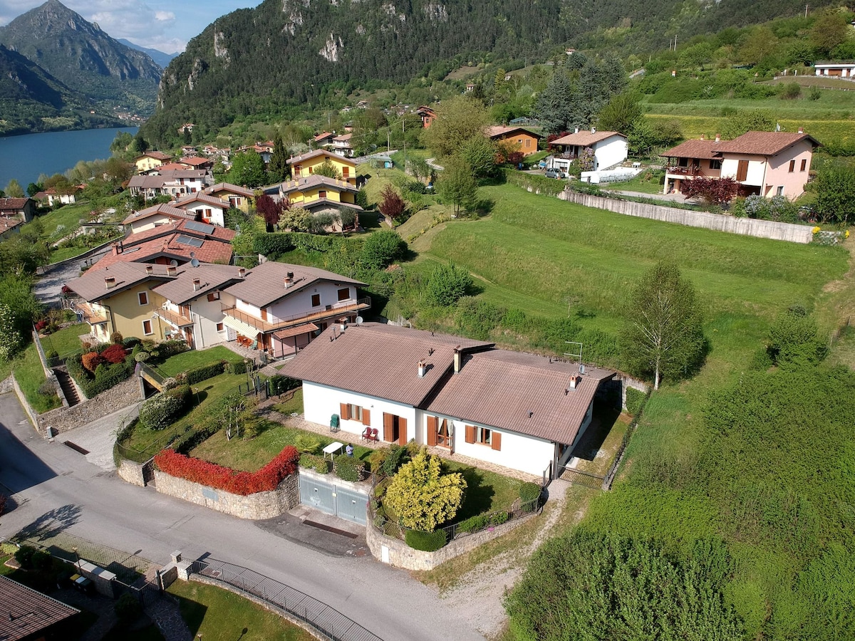 Aerial view of a tranquil countryside neighborhood featuring a cluster of homes surrounded by greenery. The serene Lake Idro is visible in the background, with mountains framing the landscape. Well-maintained gardens can be seen, enhancing the scenic environment.