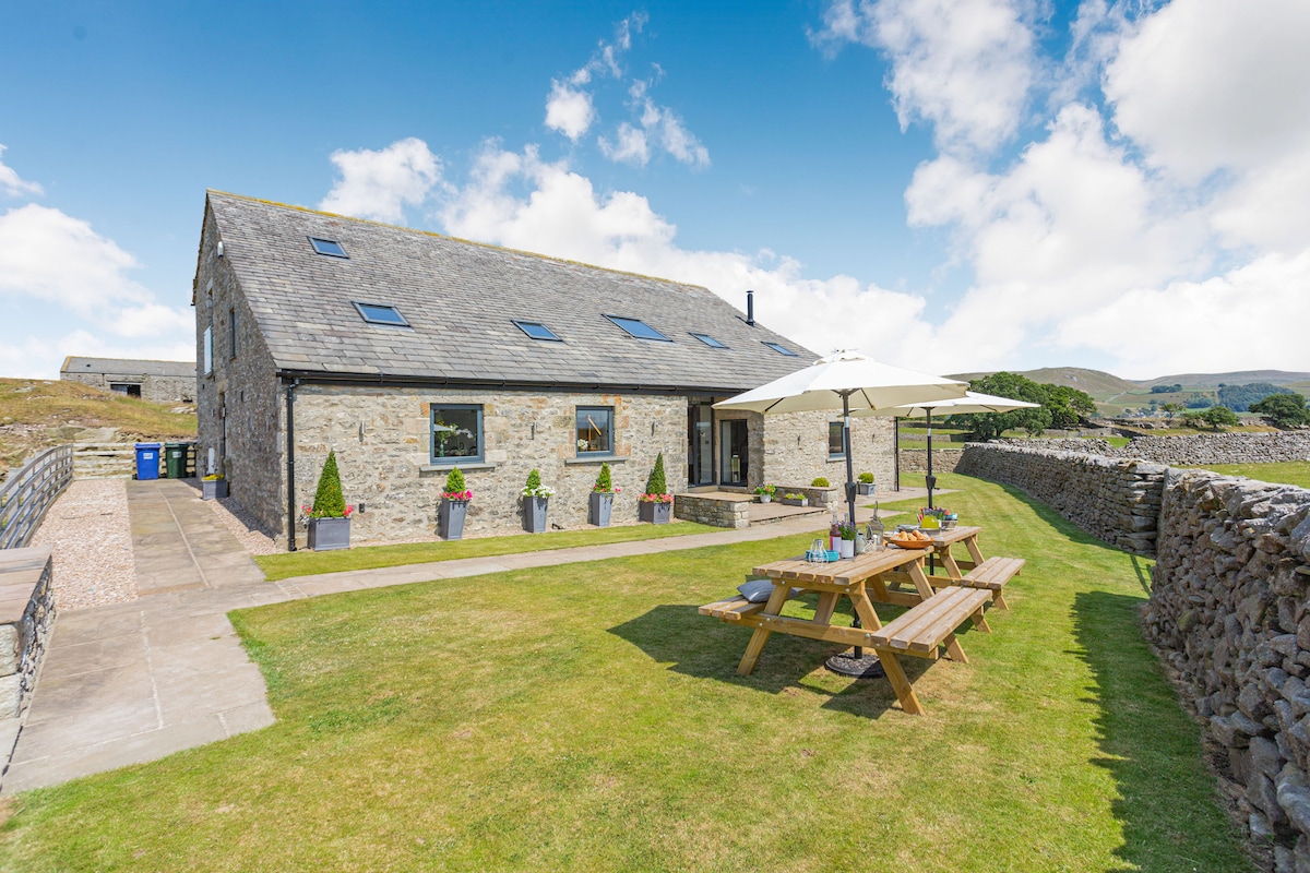 The exterior of a stone barn conversion is shown, featuring a spacious lawn and a patio area with picnic seating. Umbrellas offer shade for outdoor dining. Surrounding stone walls add to the rustic charm, while scenic countryside views can be seen in the background.