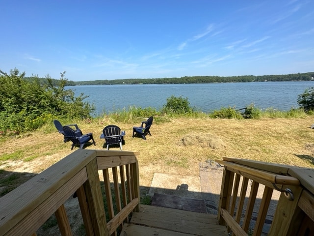 A deck leads down to a grassy area by the water's edge, where four outdoor chairs are positioned to face the calm surface of the water. Lush greenery outlines the scene, providing a tranquil setting for relaxation.