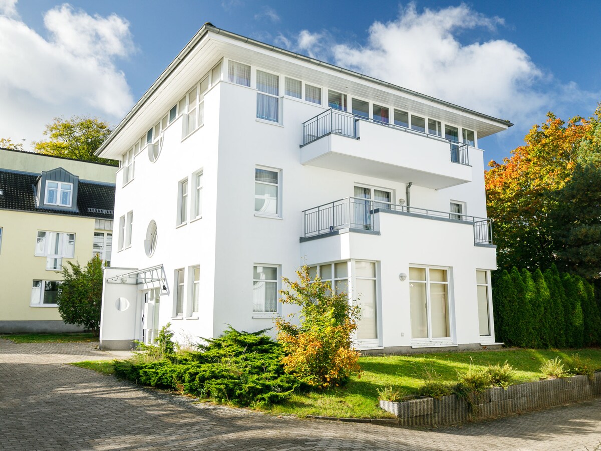 The exterior of the white three-story apartment block is highlighted by numerous windows reflecting natural light. The surrounding greenery includes neatly trimmed shrubs and grass, providing a serene atmosphere. Several balconies offer outdoor space on the upper floors, enhancing the building's inviting appearance.