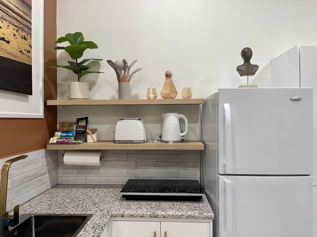 A modern kitchen area showcases a clean countertop with a sleek stove and a white refrigerator. Decorative items include potted plants, wooden utensils, and minimalist sculptures arranged on open shelves above. Granite surfaces provide a stylish contrast to the warm-colored wall.