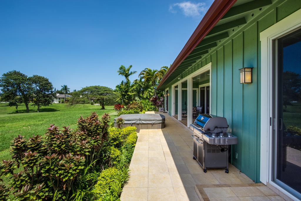 An outdoor patio features a private hot tub and a barbecue grill, surrounded by lush greenery and tropical plants. Expansive lawn areas extend beyond the patio, with distant views of palm trees and a clear blue sky overhead.