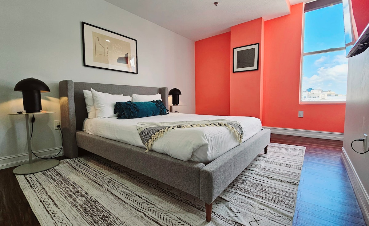 A spacious bedroom is adorned with vibrant orange and neutral walls. The room features a comfortable bed with soft linens, complemented by two stylish lamps on either side. Natural light pours in from a large window, illuminating the elegant area rug that adds warmth to the space.
