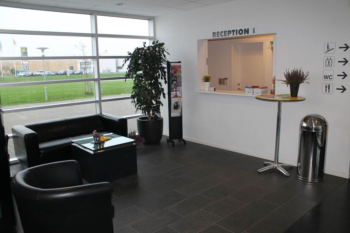 The reception area features large windows allowing natural light, complemented by black leather seating and a coffee table. A tall plant adds a touch of greenery, while directional signs guide guests to facilities. A reception desk is visible in the background.