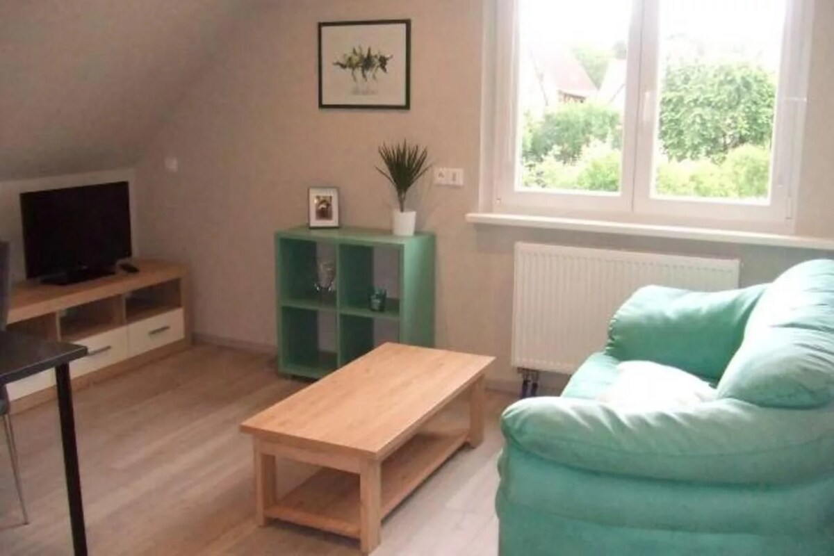 A living room area is presented with a comfortable teal sofa and a wooden coffee table. A television is positioned on a light-colored cabinet beneath a framed plant print. A large window allows natural light to fill the space, offering views of the greenery outside.