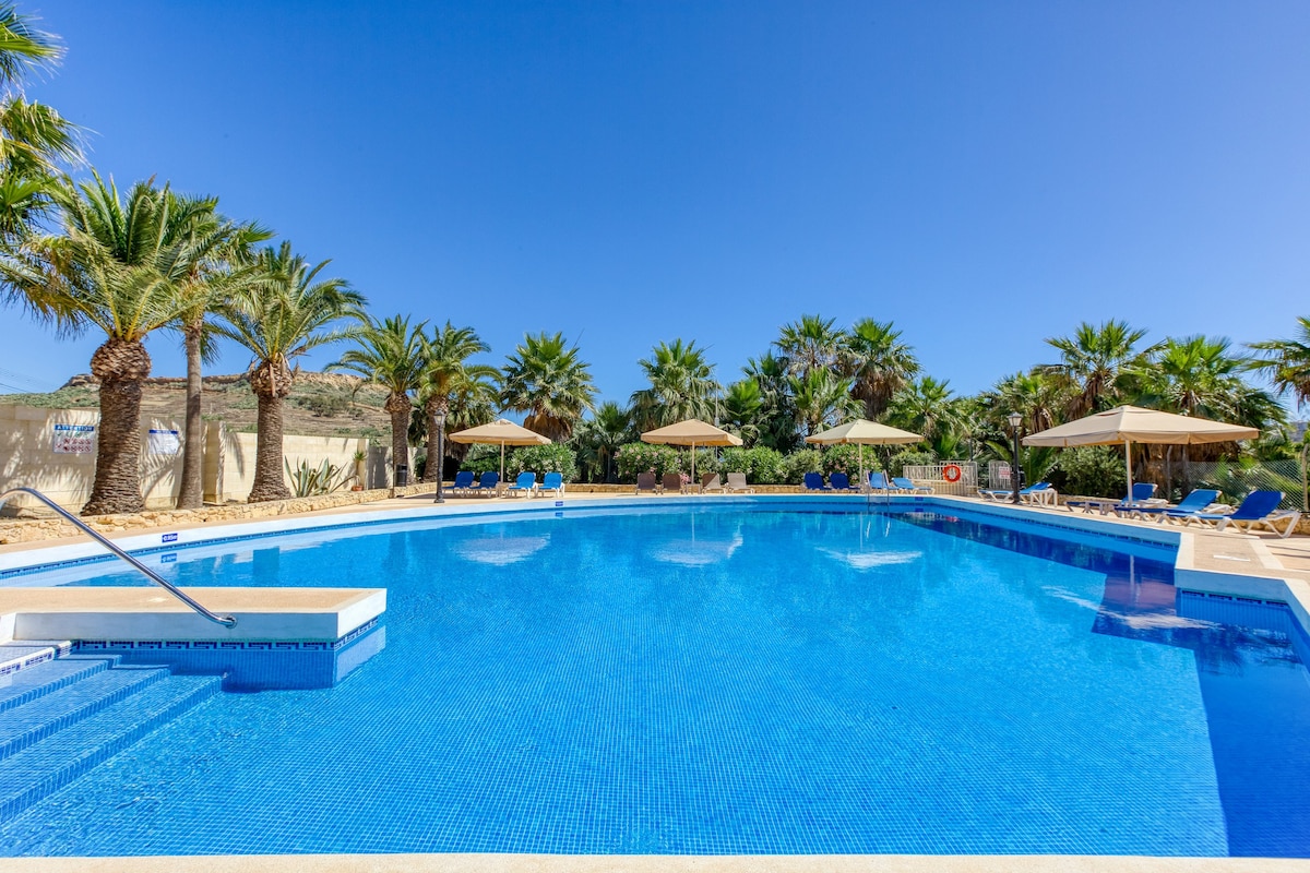A large swimming pool is surrounded by palm trees and sun umbrellas. The bright blue water reflects the clear sky above, providing a serene setting for relaxation. Lounge chairs are positioned around the pool area, ensuring ample space for comfort.