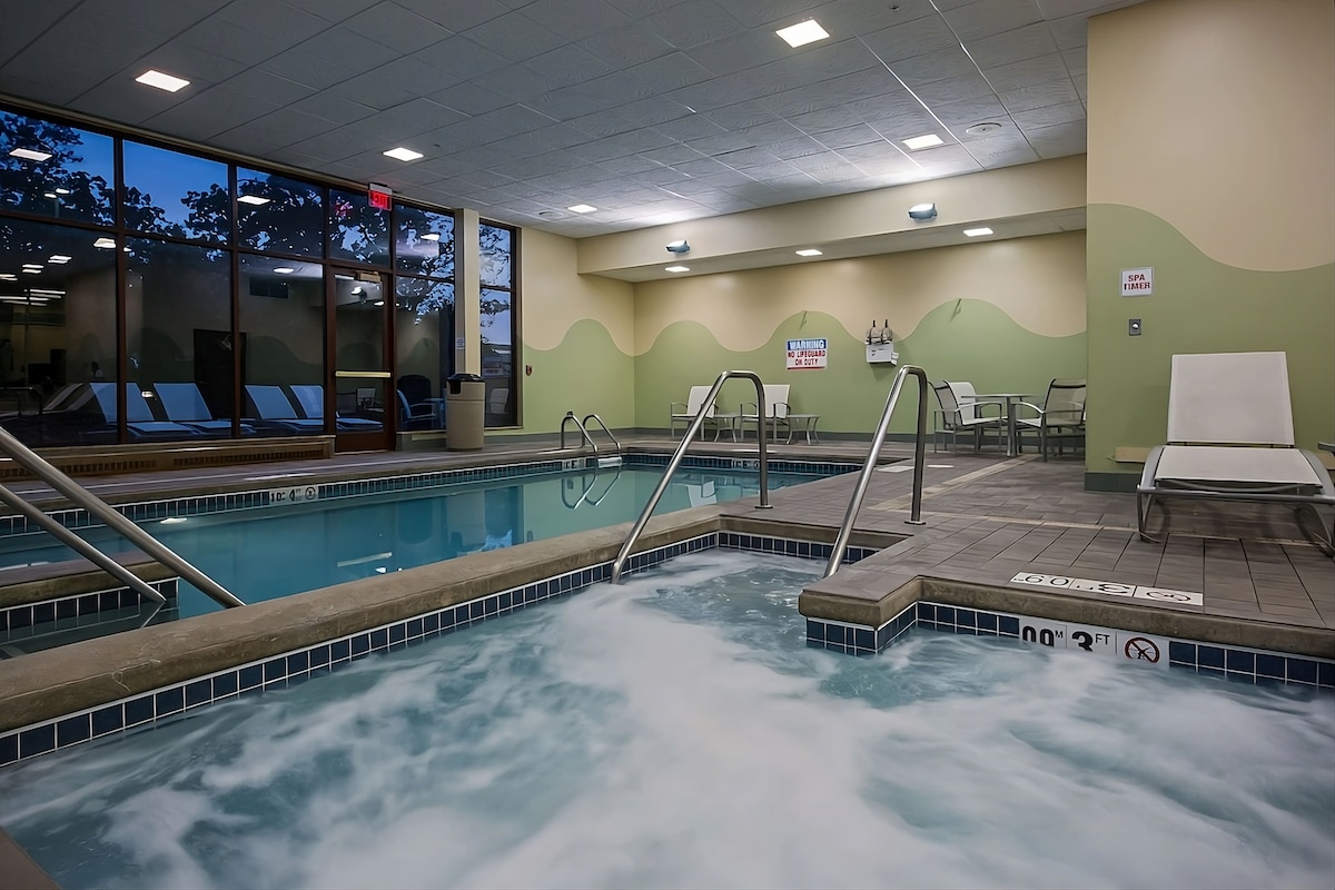 An indoor swimming pool area showcases tranquil waters with foam gently rising. Lounge chairs are positioned along the poolside, complemented by natural light filtering through large windows. Safety signage is visible, ensuring a safe environment for guests.