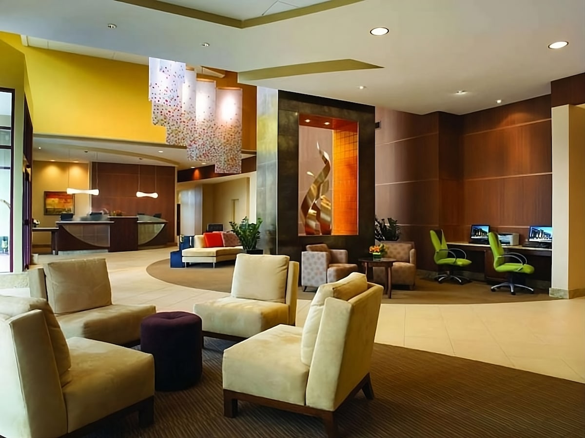 The lobby features a spacious seating area with beige lounge chairs and a purple ottoman. A colorful art installation hangs from the ceiling, and two computers are available for guest use in a bright corner. Natural light filters in, creating an open atmosphere.