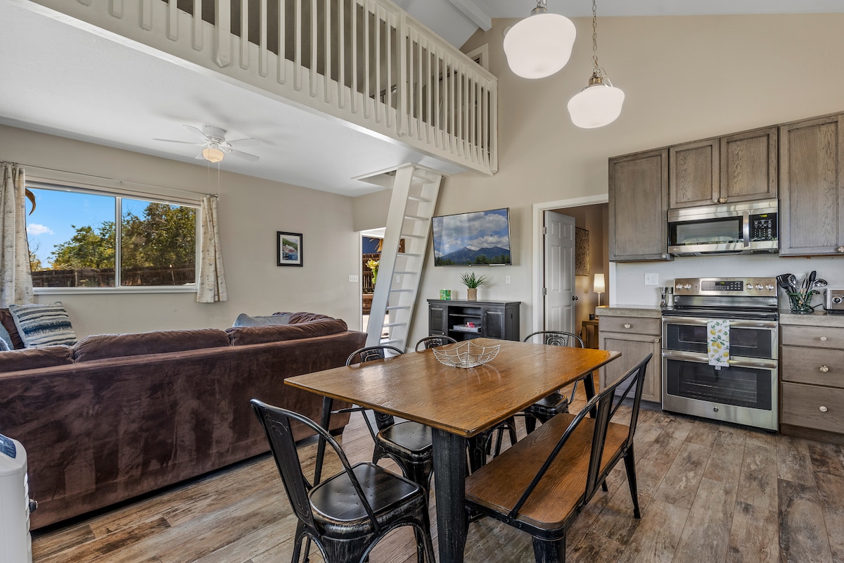 An open living area features a plush sofa and a dining table surrounded by black metal chairs. Natural light floods in through a large window, creating a spacious atmosphere. A fully equipped kitchen is visible in the background, complemented by warm wooden cabinetry.
