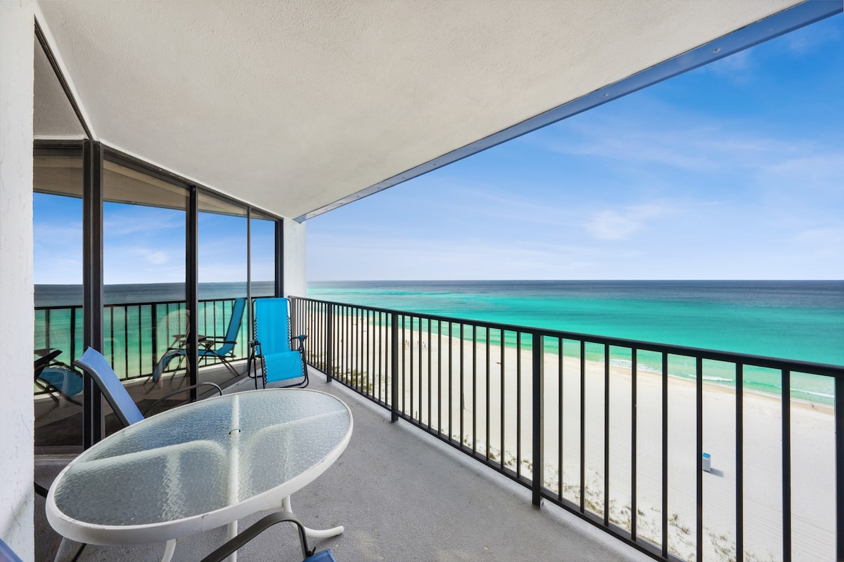 A private balcony offers unobstructed views of the beach and the Gulf of Mexico. Outdoor seating is provided by a glass table and two blue lounge chairs. The expansive ocean stretches into the horizon under a clear sky.