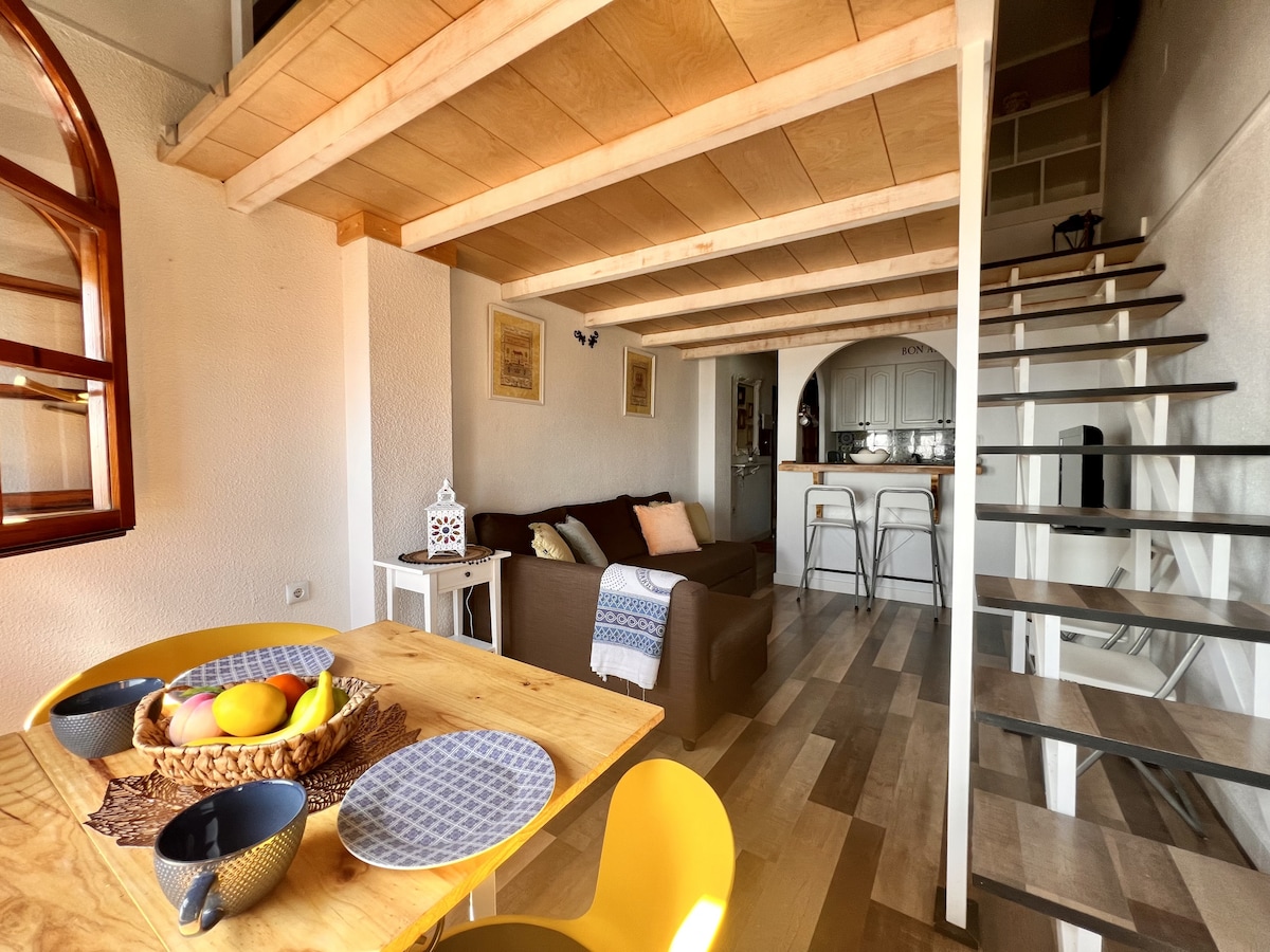 The living area features a cozy sofa and a wooden dining table set with bowls and a fruit basket. The open layout is complemented by light-colored wood flooring, and a spiral staircase leads to the upper level. Natural light filters through arched windows.