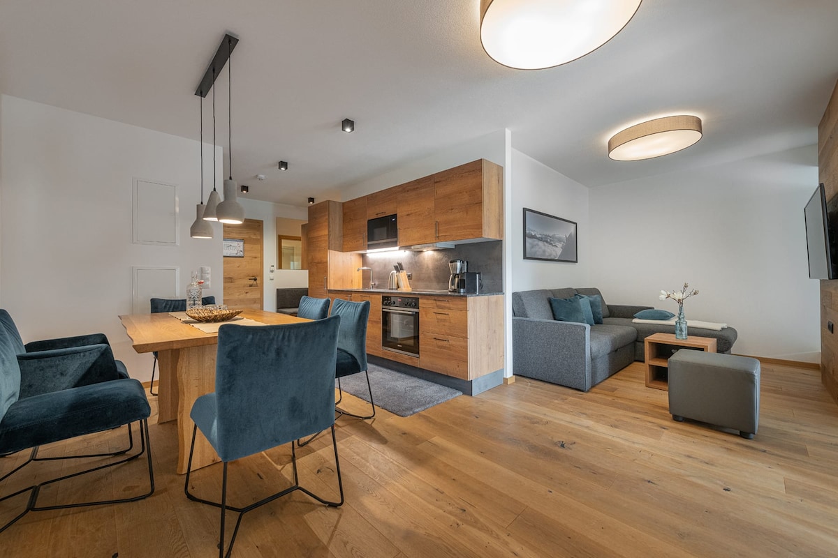 A modern living area features a dining table surrounded by four blue chairs. An open kitchen is equipped with a countertop, oven, and coffee maker. A comfortable gray sofa is positioned nearby, complemented by a small side table. Natural light enhances the warm wooden flooring.