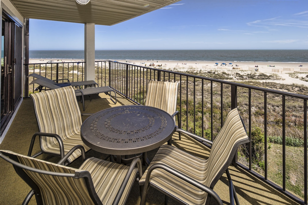 A private deck featuring a circular table surrounded by several striped lounge chairs offers stunning ocean views. The expansive sandy beach and gentle surf are visible in the distance, providing a serene backdrop for outdoor relaxation.