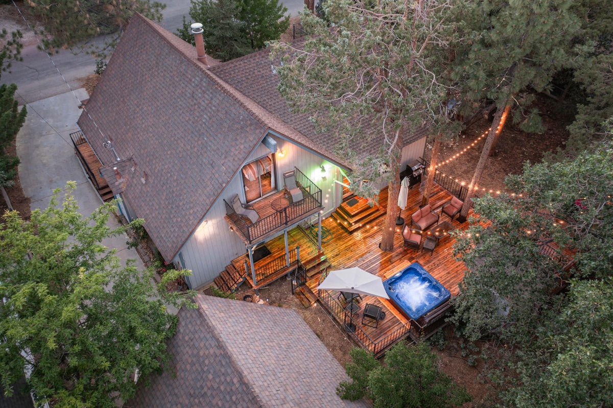 An aerial view captures the charming exterior of the home, highlighting a spacious deck with outdoor seating and a private hot tub. Surrounding trees provide a natural barrier, while soft outdoor lighting creates a welcoming atmosphere for evening relaxation.