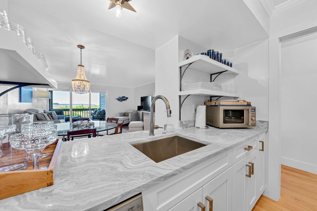 The modern kitchen space features quartzite countertops and white cabinetry, with a sink and stainless steel appliances visible. Open shelves display neatly arranged dinnerware. In the background, the living area opens up to a deck with ocean views, enhancing the room's bright and airy feel.