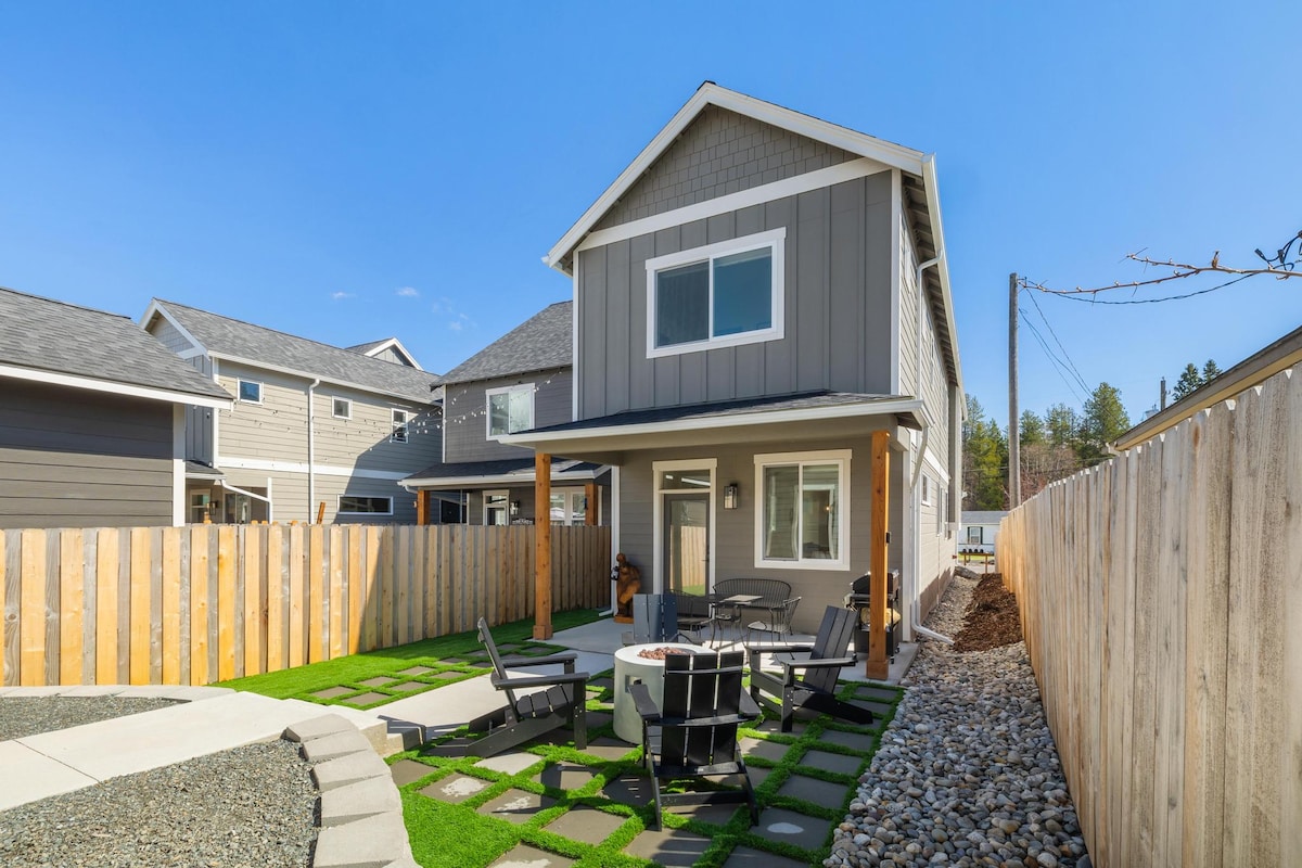 A modern two-story home is set within a private, fenced yard. Outdoor seating is arranged around a fire pit on a stone patio. The landscaping features decorative stones and green grass, complemented by wooden privacy fencing and clear blue skies.