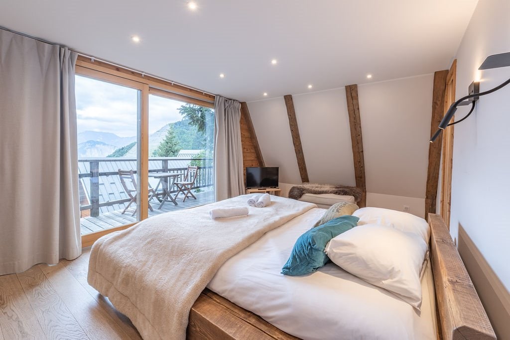 A master bedroom features a spacious bed with a light-colored blanket and decorative pillows. Large windows allow natural light to enter, revealing a balcony with outdoor seating and mountain views. Soft curtains frame the windows, providing a cozy ambiance.