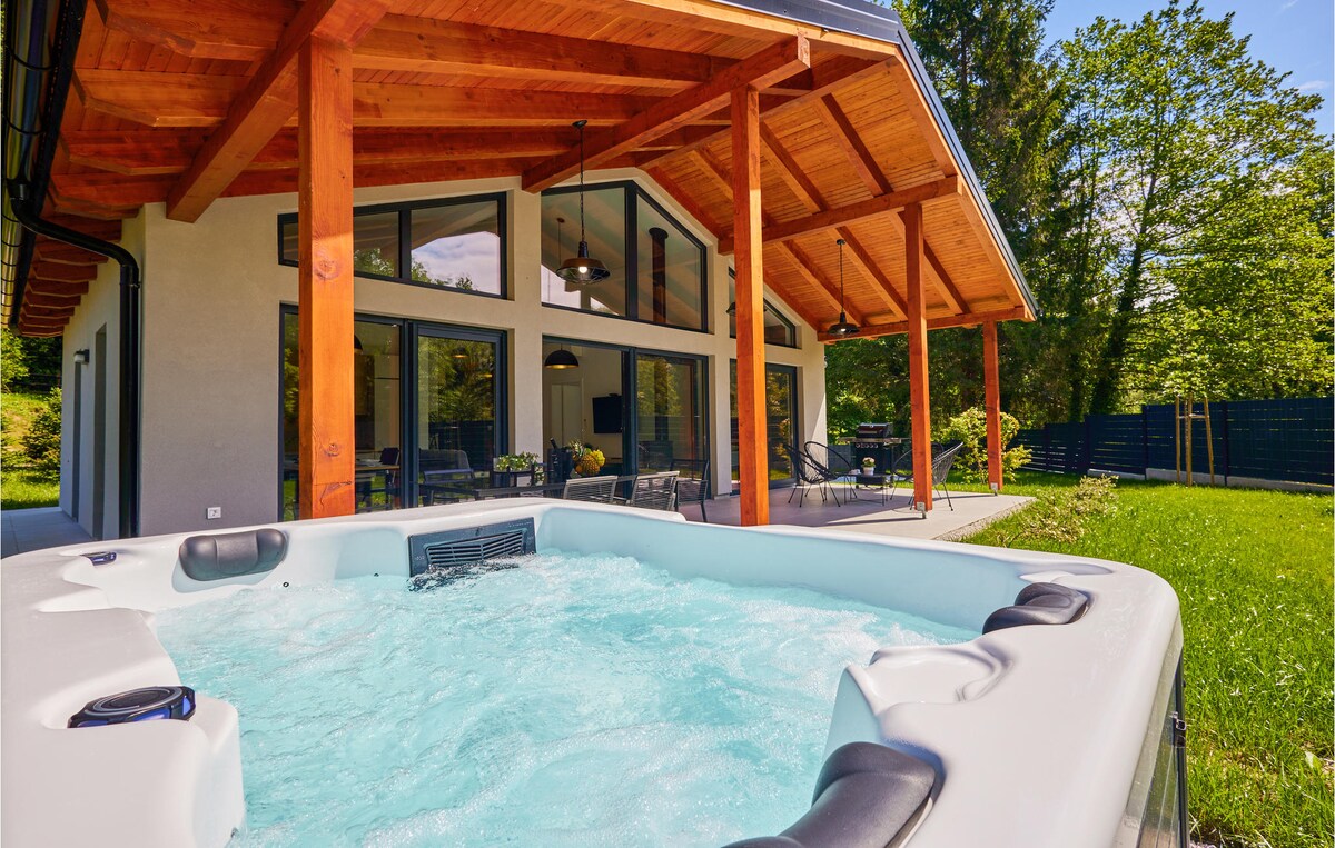 A spacious whirlpool is positioned in the foreground, surrounded by a well-kept lawn. The modern holiday home features large glass windows under a wooden roof, allowing natural light to illuminate the interior. Lush greenery encircles the property, enhancing the serene outdoor environment.