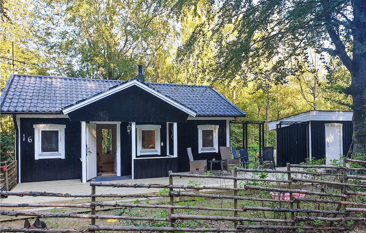 A black wooden cabin surrounded by trees is shown, featuring a sloped roof and a welcoming front porch. Outdoor seating areas are visible, alongside a separate structure. The natural setting indicates a serene environment, ideal for relaxation and recreation.