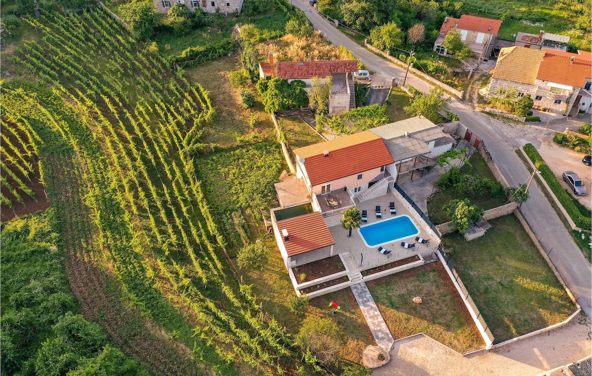 An aerial view of the vacation home reveals a private swimming pool surrounded by a spacious patio. Lush green vineyards fill the landscape, with residential buildings and a road visible nearby. The red-tiled roof of the home contrasts with the greenery.