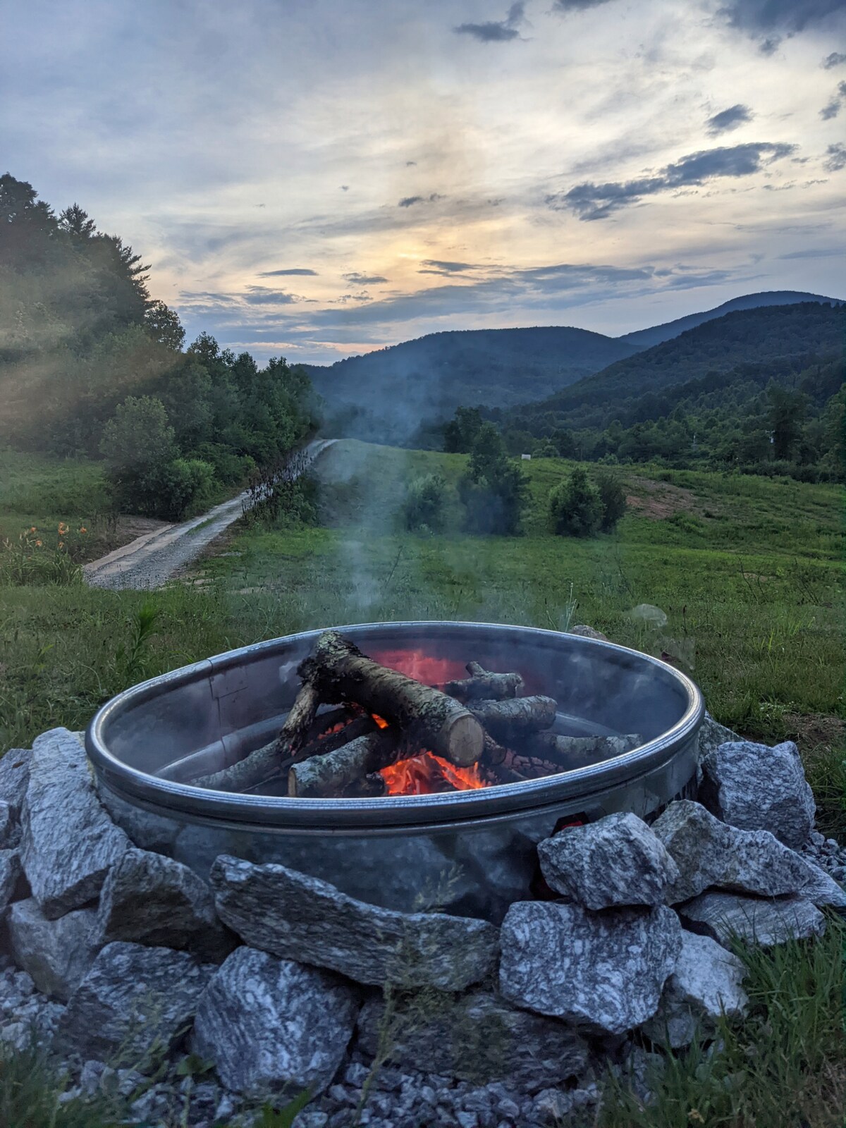A fire pit is surrounded by a circle of stones, with logs burning in vibrant flames. The backdrop features rolling green hills and mountains under a sky transitioning into evening, with wisps of clouds creating a serene atmosphere.