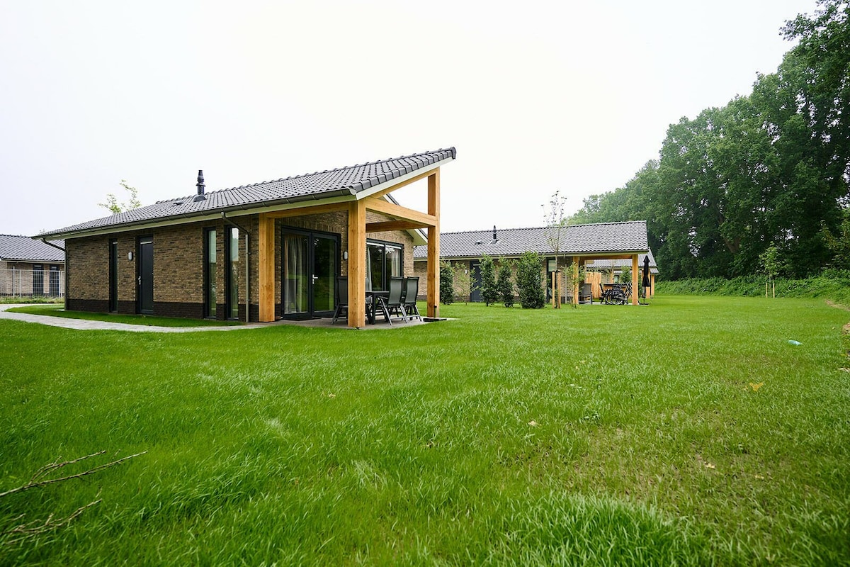 The villa features a modern exterior with a combination of wooden beams and dark brick. Lush green grass surrounds the building, which includes large windows reflecting the outdoor scenery. A covered terrace with seating is visible, offering a seamless connection to the natural surroundings.
