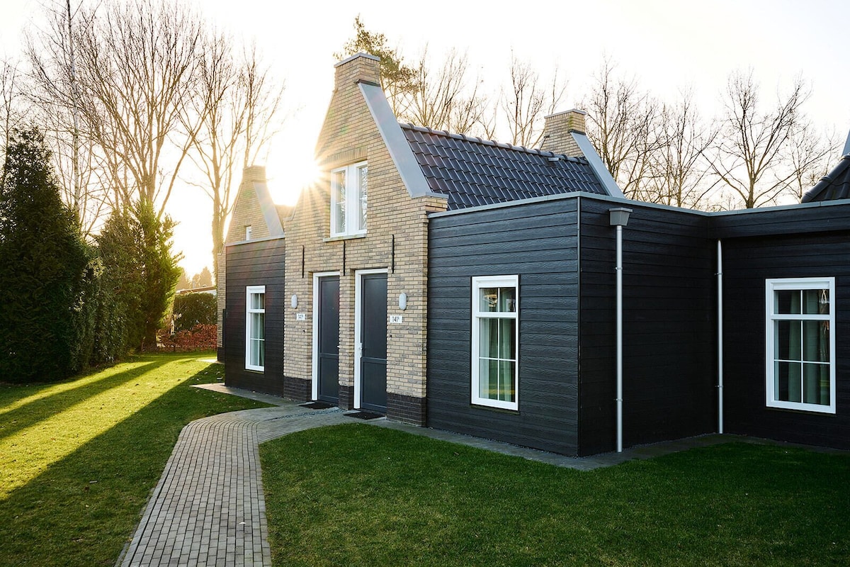 A semi-detached villa is surrounded by a well-maintained lawn, with a winding pathway leading to the entrance. Sunlight filters through the trees, highlighting the modern architectural details of the exterior, which features a mix of dark siding and brick elements.