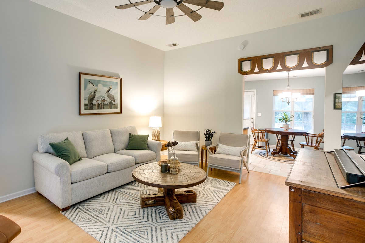 A welcoming living area is offered with a light-colored sofa and armchairs surrounding a round wooden coffee table. A ceiling fan hangs above, while natural light fills the space, highlighting the neutral tones and simple decor. A dining area can be seen in the background.