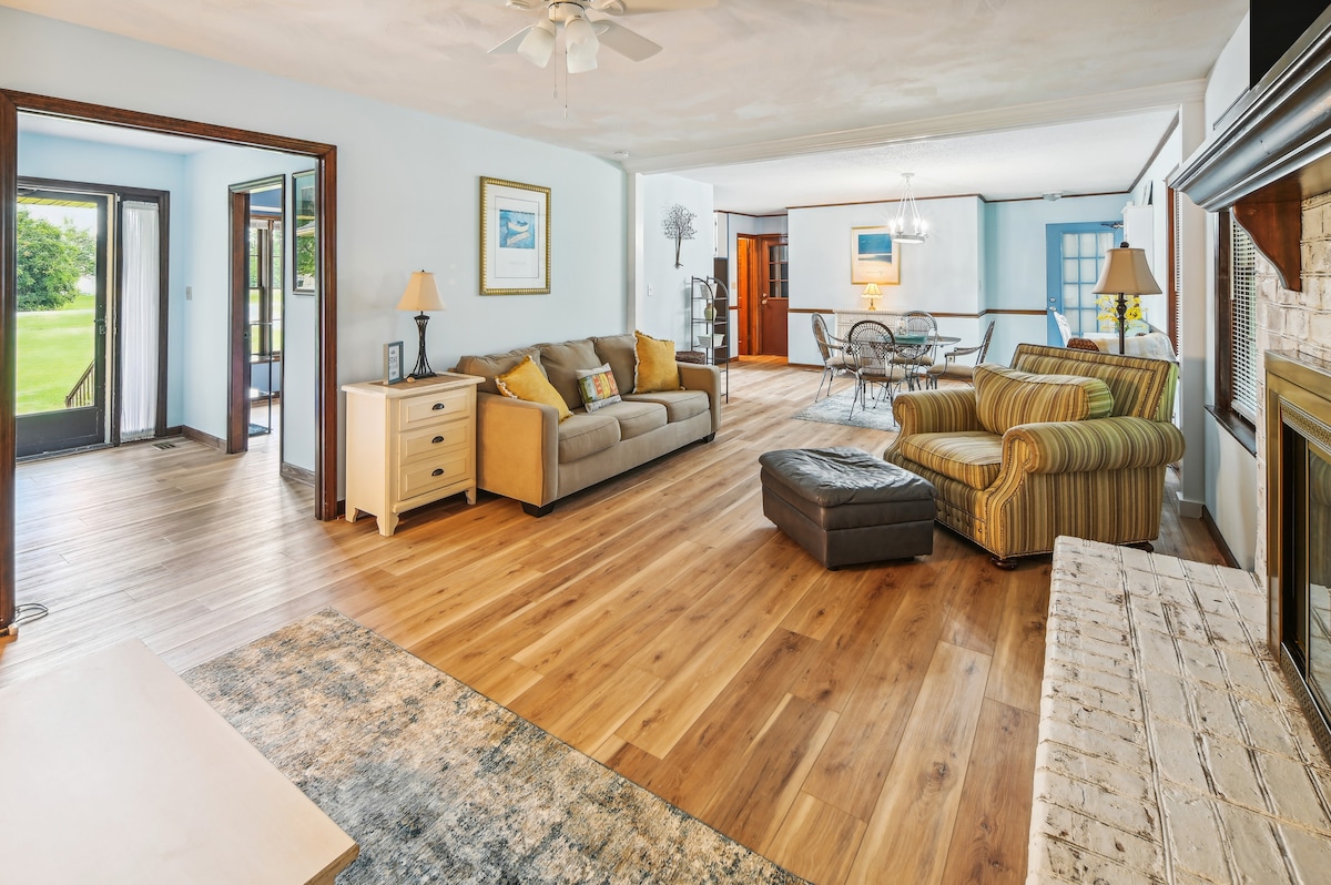 A spacious living area features a combination of light wood flooring and neutral walls. Soft seating options include a sofa and striped chair, while a decorative area rug adds texture. Natural light streams through large windows, providing views of the adjoining outdoor space.