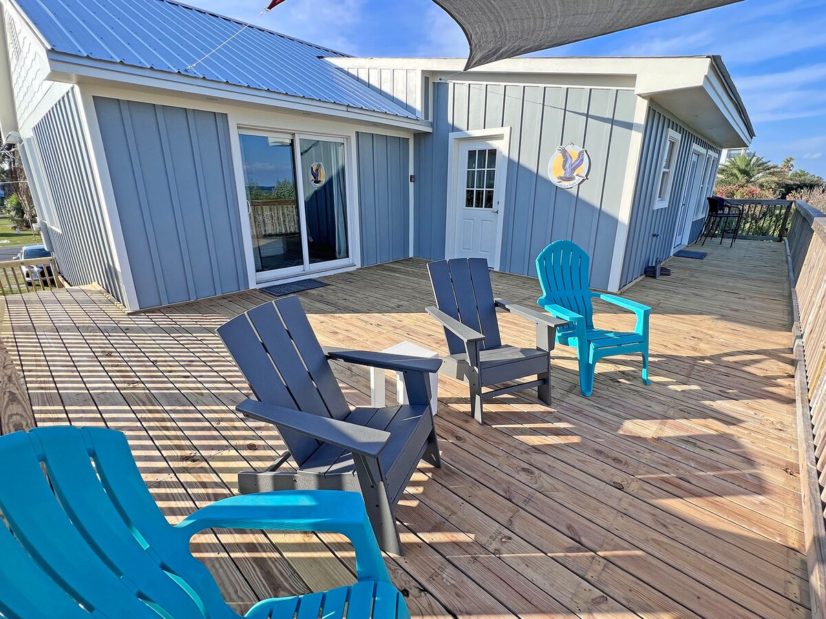 A spacious deck features a mix of blue and black Adirondack chairs arranged for relaxation. The light wood decking is complemented by a nearby entrance and large sliding glass doors, allowing easy access to the indoor living space.