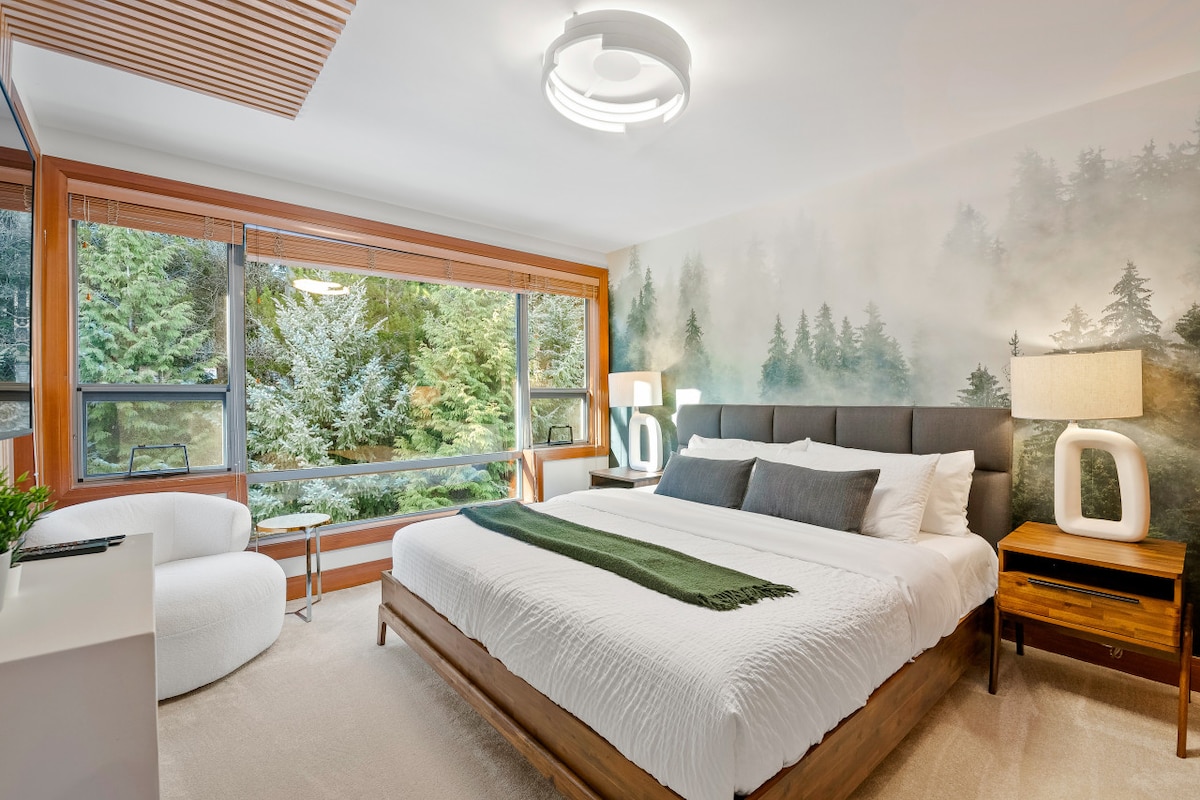 A serene bedroom features a spacious bed with a soft white blanket and dark headboard. Large windows provide a view of lush greenery, allowing natural light to brighten the room. Modern lighting fixtures and neutral decor create a relaxing environment.