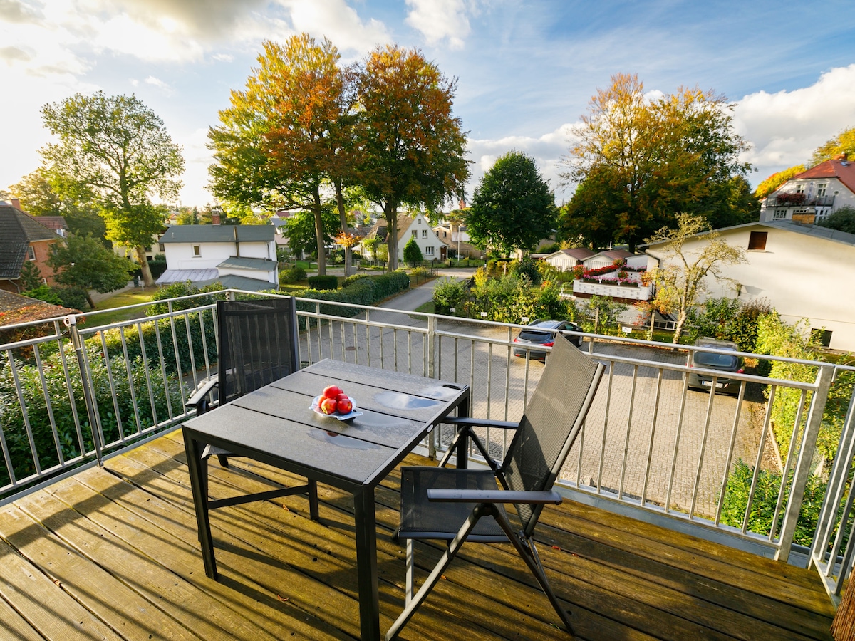 A balcony view is presented with a wooden deck, featuring a black table and two chairs. A bowl of red fruits is placed at the center of the table. Surrounding the area, trees show autumn colors, and a residential street is visible.