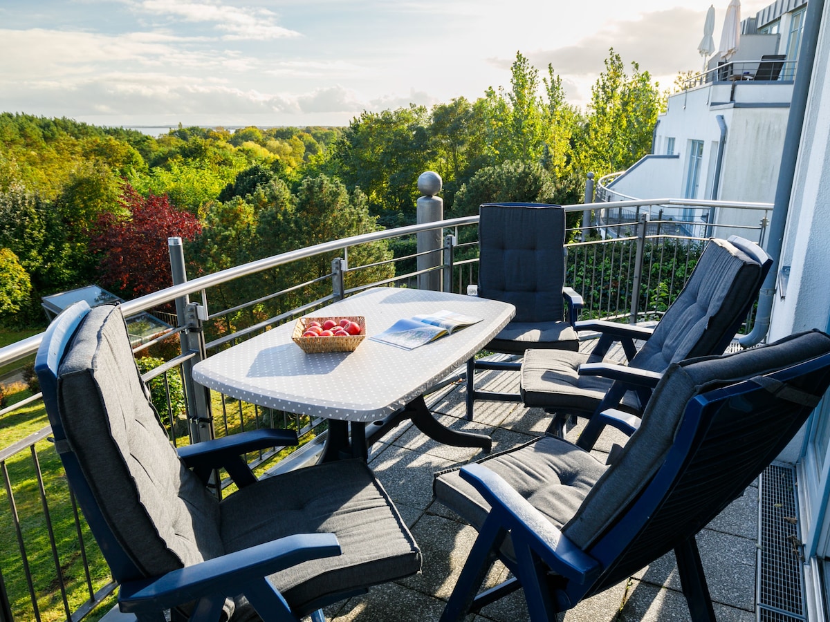 A balcony space features four cushioned chairs surrounding a table. A fruit basket sits on the table, and lush greenery is visible in the background, creating a peaceful outdoor setting.
