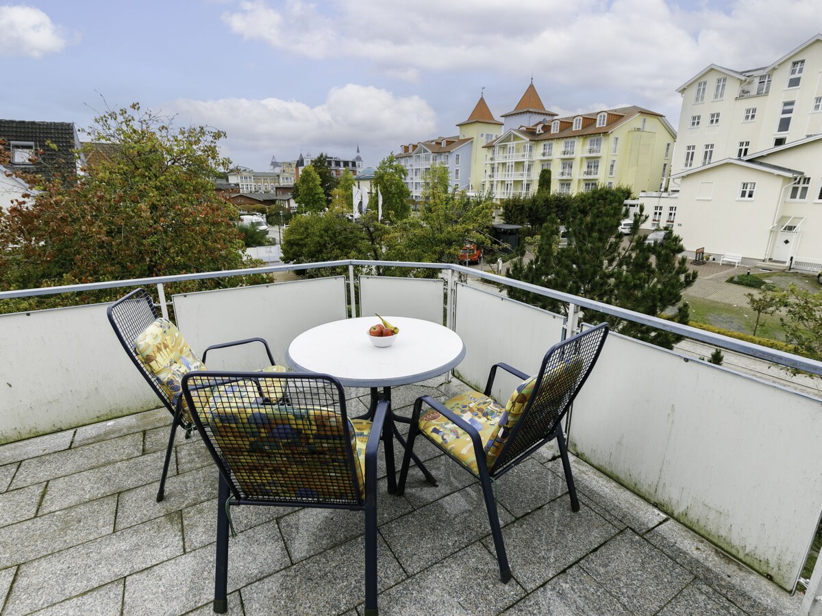 A balcony area features a round table accompanied by four chairs with colorful cushions. The surrounding view includes buildings and greenery, with cloudy skies overhead. The balcony is enclosed by a railing, providing an open and inviting space for relaxation.