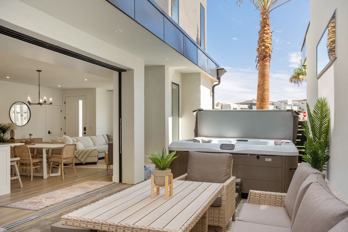 The outdoor space features a comfortable seating arrangement with a light-colored table and soft cushions. A private hot tub is positioned nearby, offering relaxation options. Large sliding doors lead to an interior space, framed by a palm tree, enhancing the tropical ambiance.