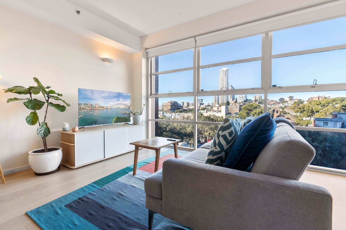 A bright living area is filled with natural light, featuring floor-to-ceiling windows that offer expansive city views. A neutral-toned sofa is paired with a colorful area rug and a wooden coffee table. A decorative plant enhances the space, with a modern TV unit positioned against the wall.