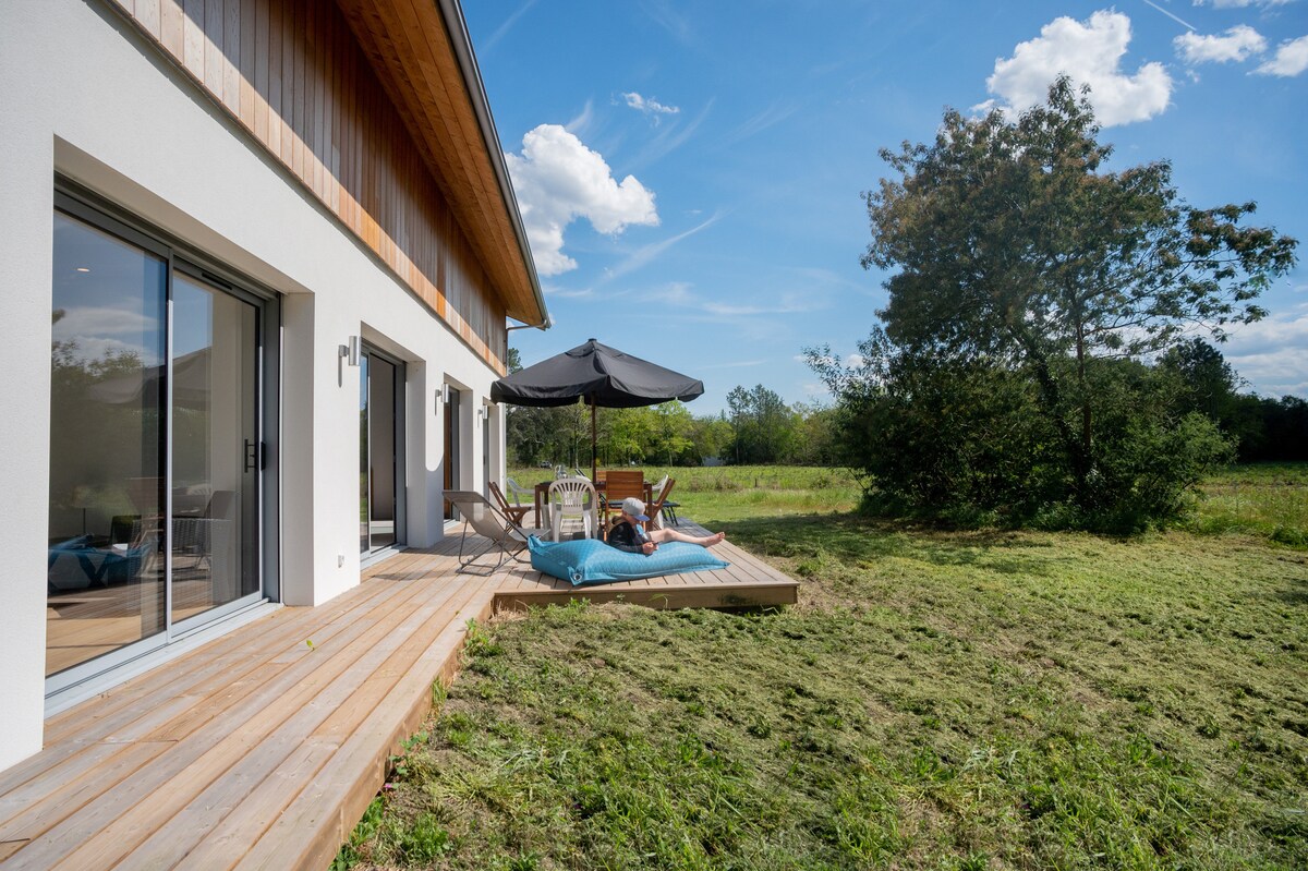 A sunlit terrace is equipped with a dining table under an umbrella, accompanied by chairs and a lounge area featuring a bright blue blanket. The wooden deck extends into a grassy area, surrounded by trees and under a clear blue sky dotted with clouds.