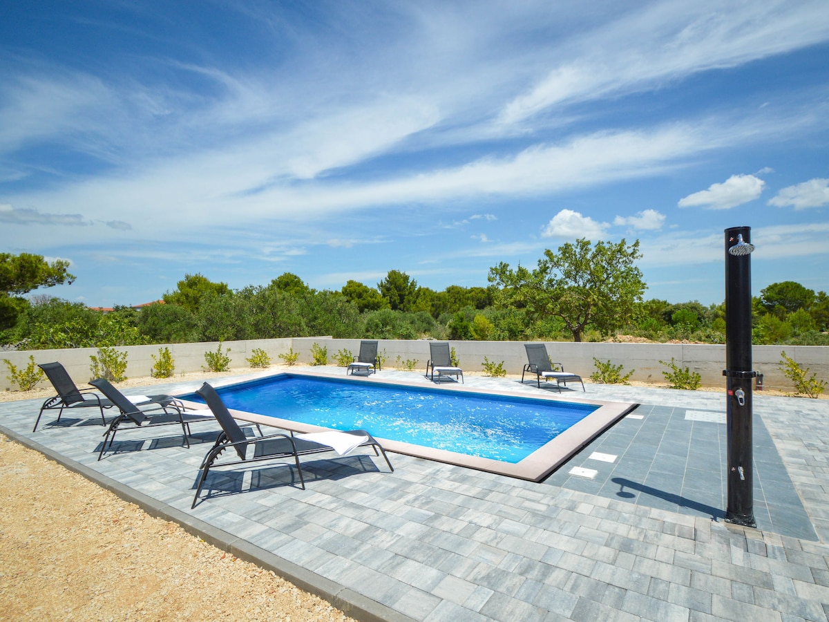 An angular swimming pool is surrounded by a stone patio and equipped with lounge chairs. A dark outdoor shower is positioned nearby, with a backdrop of lush greenery and a clear blue sky, creating a serene environment for relaxation.