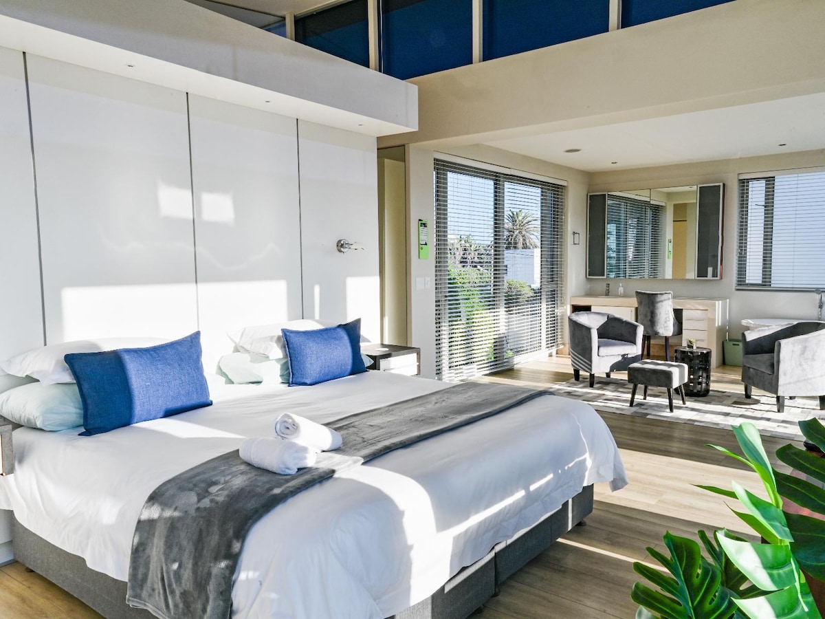 A modern bedroom features a plush king-size bed with fresh linens and decorative pillows. Natural light streams in through large windows, illuminating a cozy lounge area with comfortable chairs and a mirror. A soft grey throw is draped over the bed, enhancing the contemporary design.