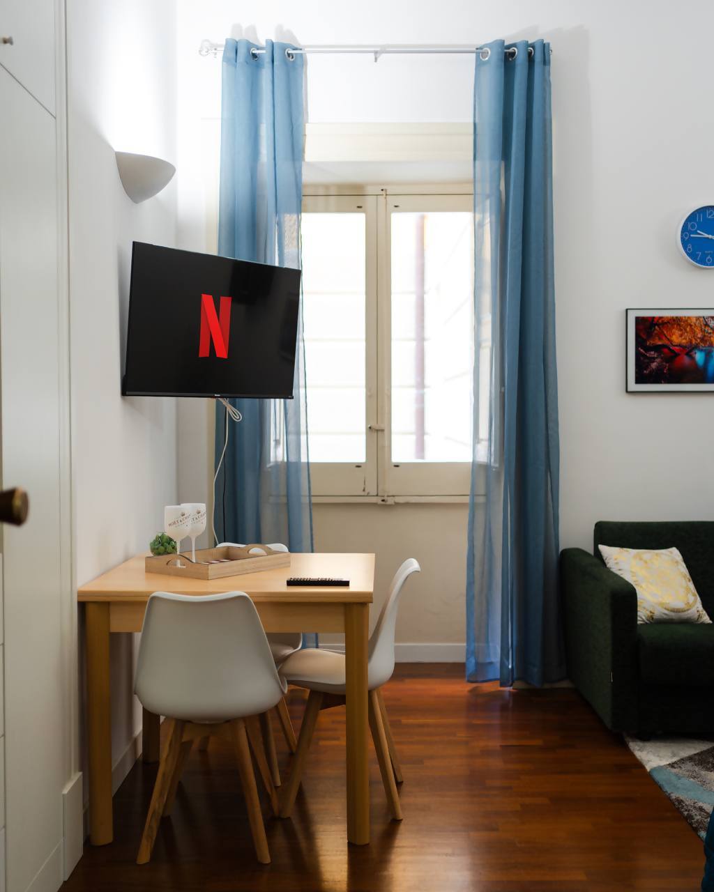 A light-filled corner of the living area is showcased, featuring a Smart TV mounted on the wall and a small dining table with two chairs. Sheer blue curtains frame a window, allowing natural light to softly illuminate the warm wooden floor.