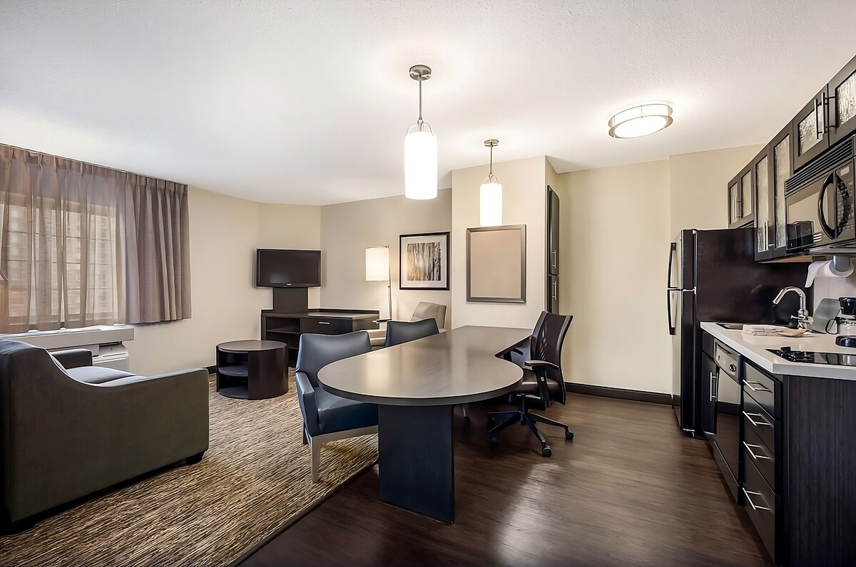 A spacious suite features a comfortable living area with a sofa and a flat-screen TV. A circular dining table is positioned near a fully-equipped kitchenette, which includes modern appliances. Soft, natural light filters through sheer curtains, highlighting the warm tones of the decor.