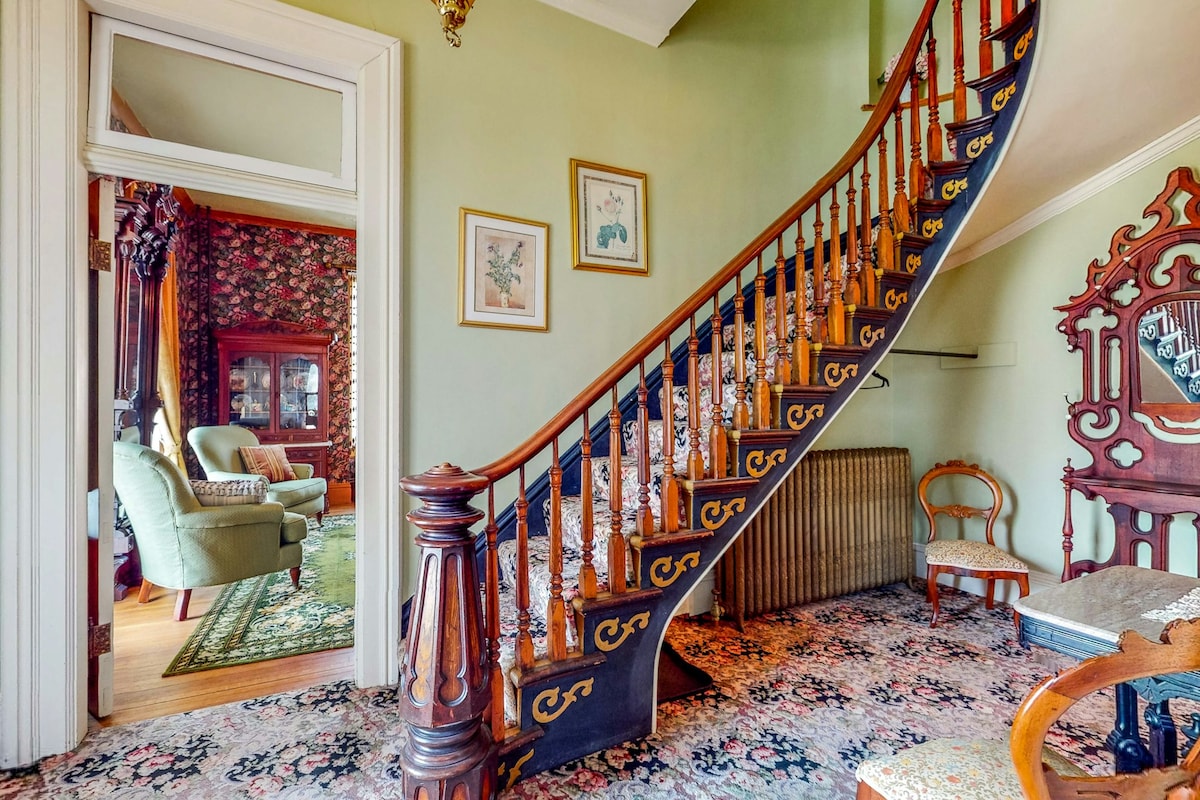 A grand staircase with elegant wooden railings leads to the upper level, flanked by a floral wallpaper and vintage furnishings. The entryway features intricate carpet patterns, adding to the historical charm of the home. An antique mirror and chair are visible in the entry space.