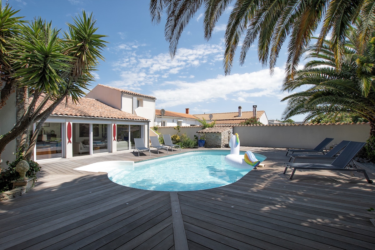 A private outdoor area features a heated swimming pool surrounded by wooden decking. Sun loungers are positioned nearby, while lush palm trees provide shade. A colorful inflatable float is present in the pool, and sliding glass doors lead to a spacious indoor area.