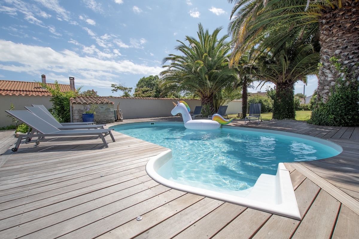 A private swimming pool is surrounded by a wooden deck and tropical palm trees. A white unicorn float is placed in the clear water. Sun loungers are positioned nearby, inviting relaxation under a sunny sky.