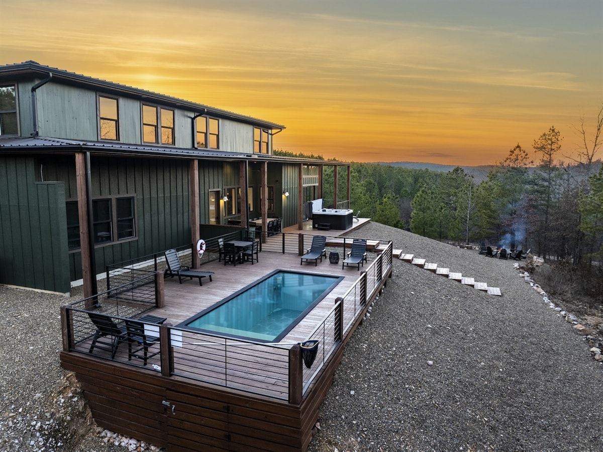 An expansive deck features a container pool surrounded by seating areas. The cabin, designed with large windows, is complemented by a backdrop of lush trees. A grill and outdoor space are visible, highlighting the seamless connection between indoor and outdoor living.
